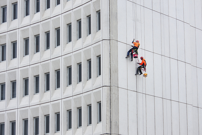 workers in orange jackets scaling a grey brick building - nice us