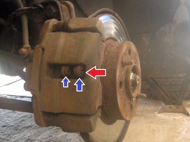 Красной стрелкой указано отверстие в скобе тормозного суппорта, а двумя синими тормозные колодки